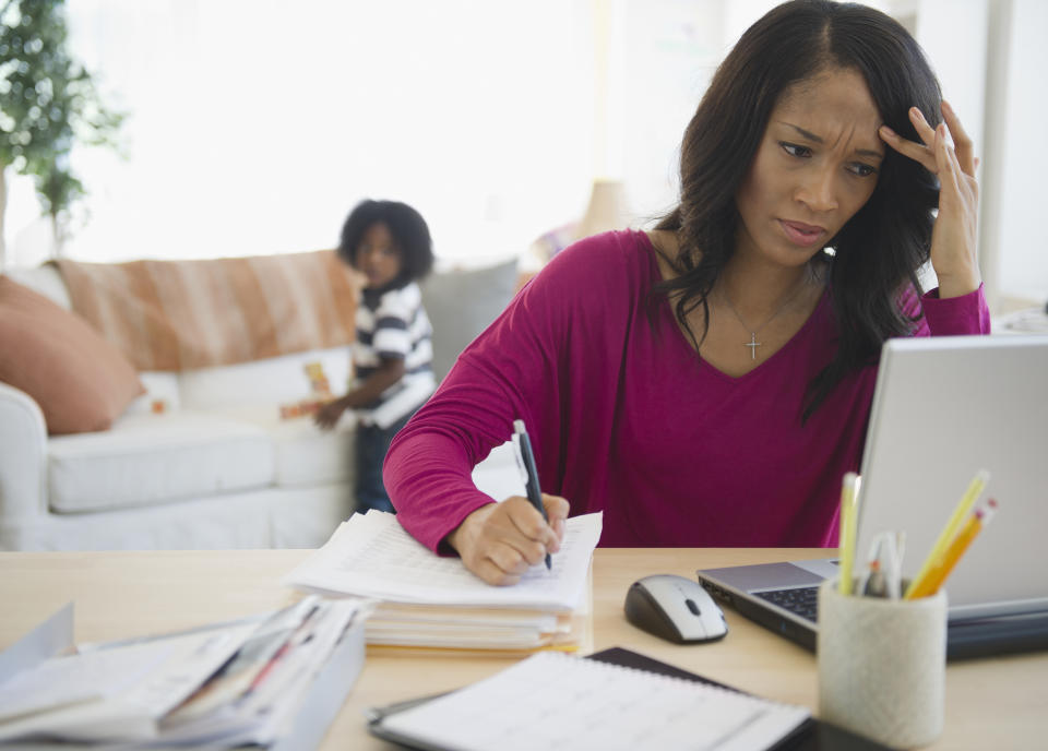 A mother looks concerned about money while looking at a laptop with her child in the background.