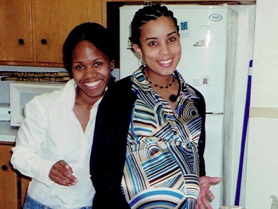 Photo of Sibylla Nash with her friend, standing in a kitchen and smiling. Sibylla has black hair braided into cornrows, brown eyes, and is pregnant. She wears a black cardigan and wears a silky collared dress that has a stripy blue, white, black, and tan pattern. She is buttering a plate full of bread. Her friend to her left has black hair tied back into a ponytail, brown eyes, and wears a white button-down shirt.