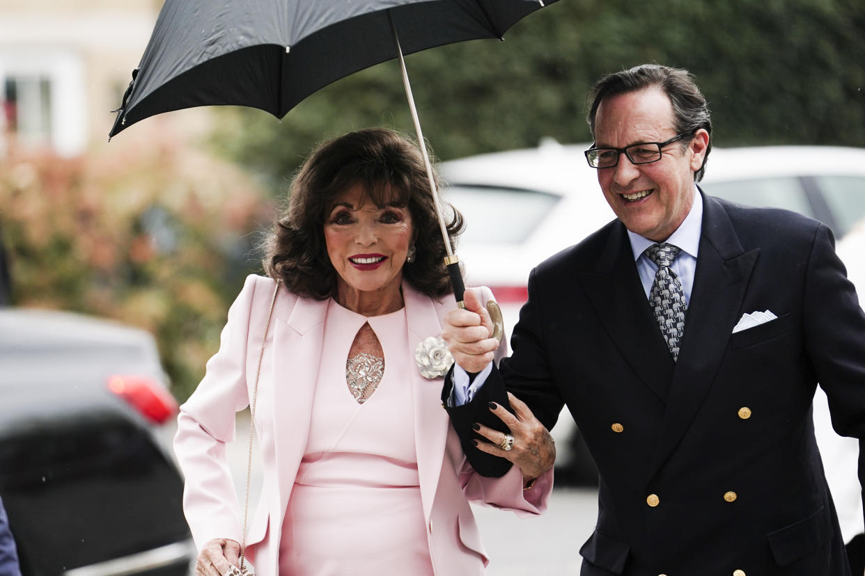 Dame Joan Collins, wearing a pink dress with a matching pink blazer, under an umbrella held by her husband Percy Gibson, wearing a black double-breasted suit, white shirt and patterned tie