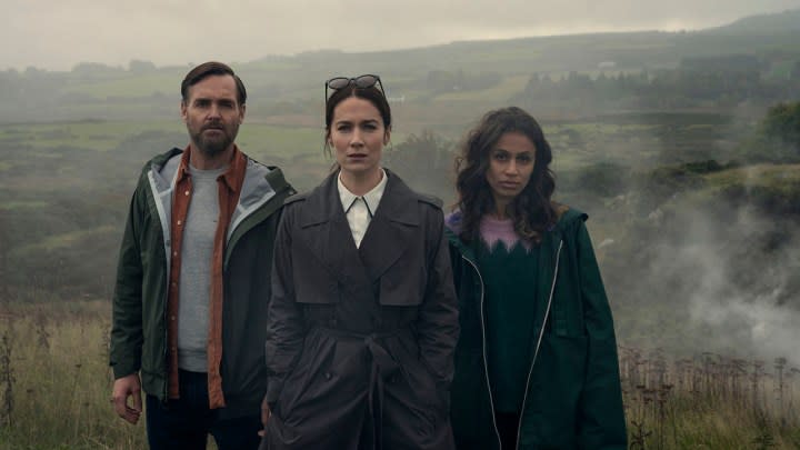 Three people standing outside on a cloudy day looking at something in a scene from Bodkin.