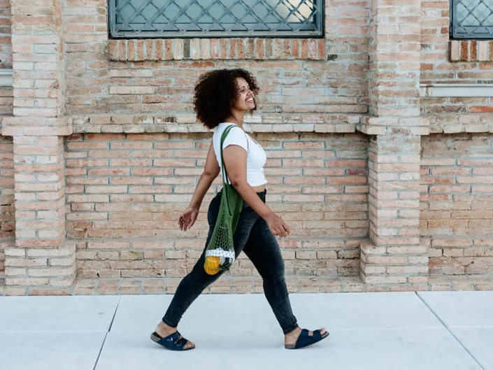 a person with dark curly hair wearing black pants and a white T shirt, walking on a brightly lit sidewalk in front of a stone building during the day