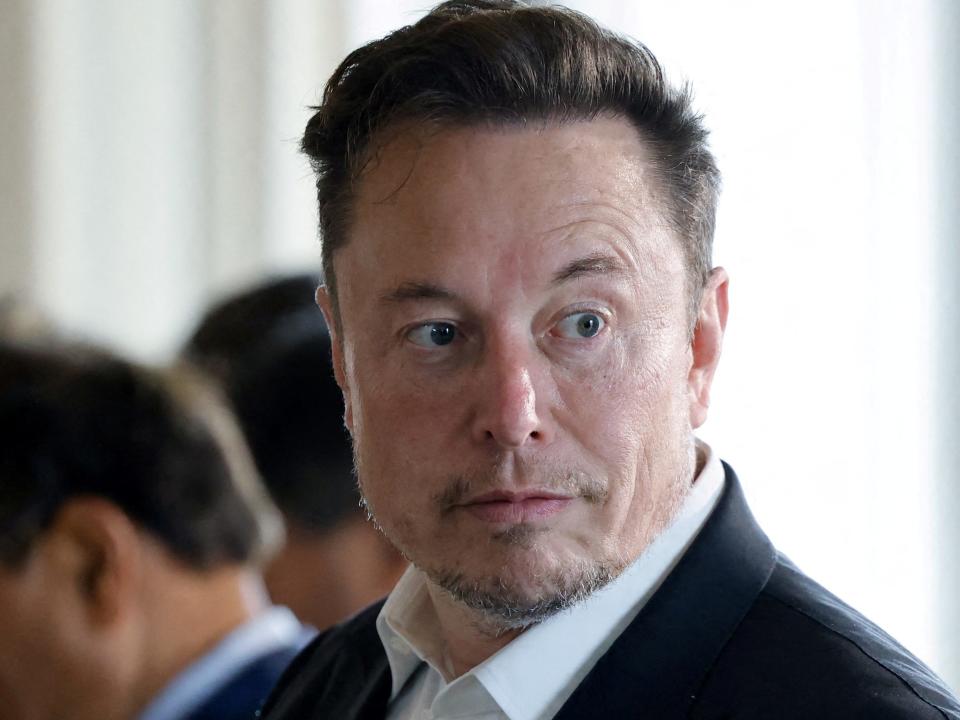 elon musk raises one eyebrow wearing a suit in as room full of seated men