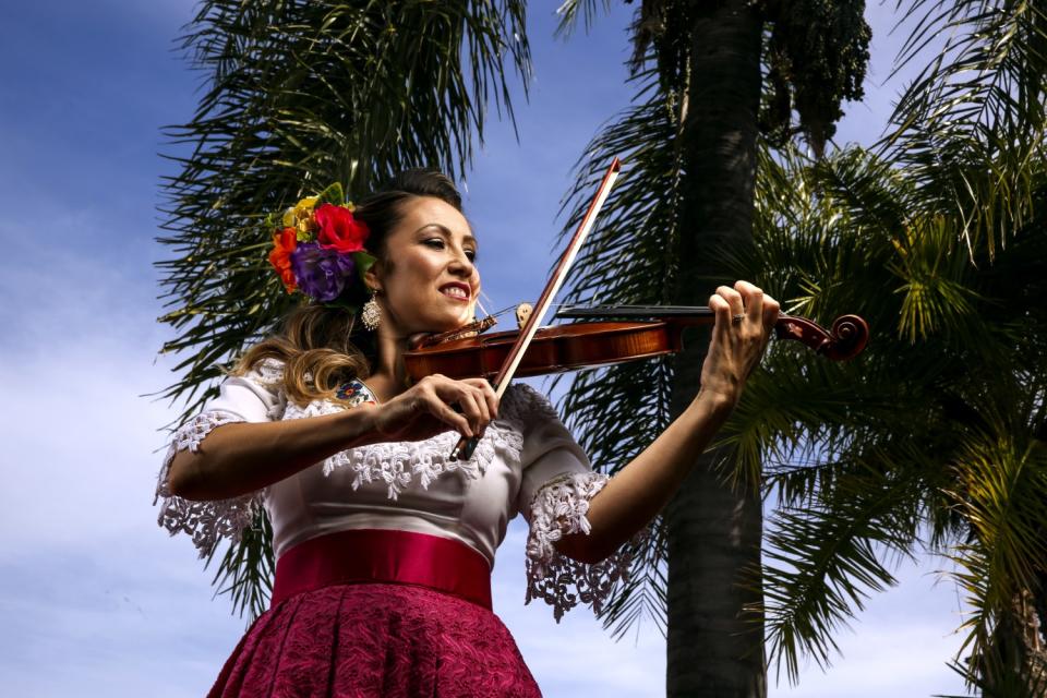 A woman holds a violin in front of palm trees