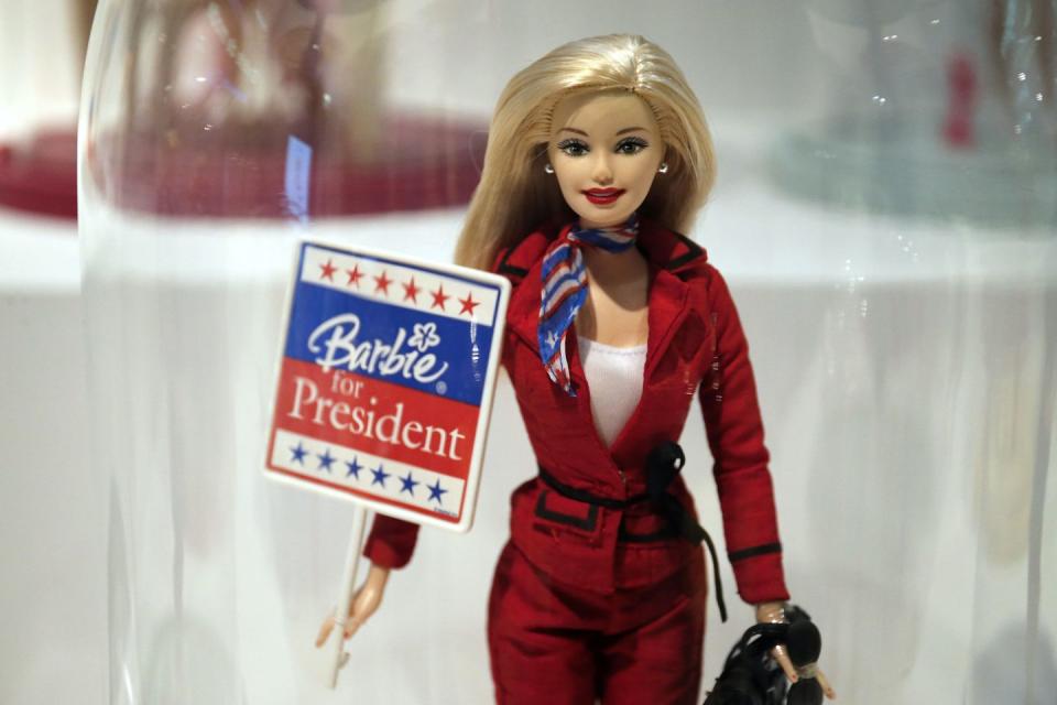 a barbie doll wearing a red pantsuit while holding a barbie for president sign