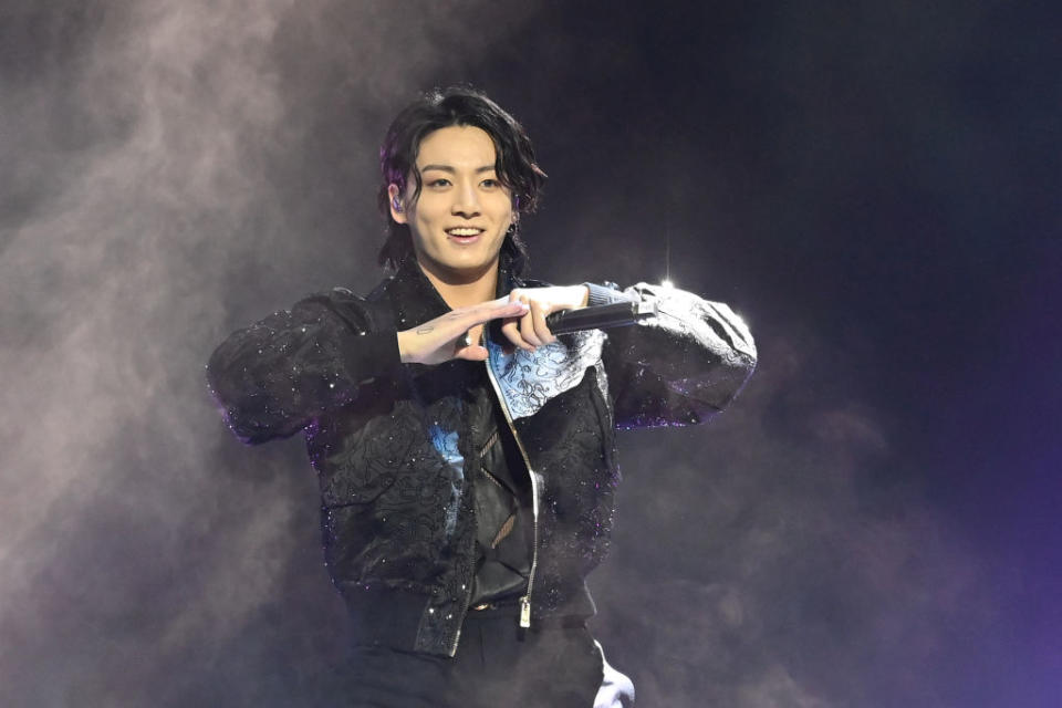 Jung Kook performs on stage, wearing a stylish black jacket, holding a microphone, making a heart shape with his hands. He is smiling and surrounded by smoke