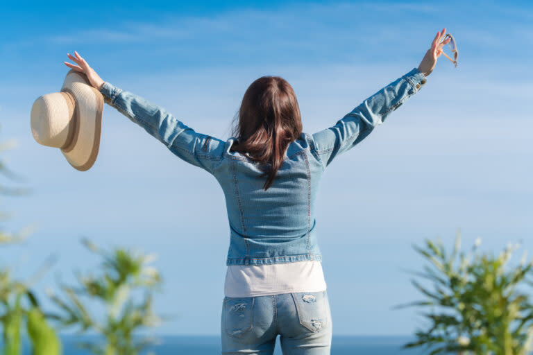 Female raising hands enjoying vacation, standing on background of blue cloudless sky.