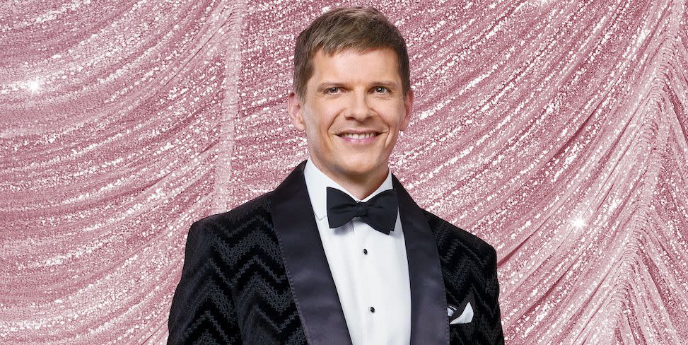 nigel harman for strictly come dancing 2023, a man stands with hands in pockets looking at the camera and smiling, he wears a black suit with bow tie and is surrounded by glitterballs
