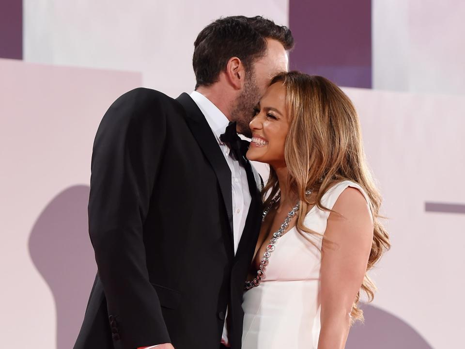 Ben Affleck, wearing a classic black tuxedo, and Jennifer Lopez, wearing a floor-length white mermaid gown, smile as they pose on a red carpet.