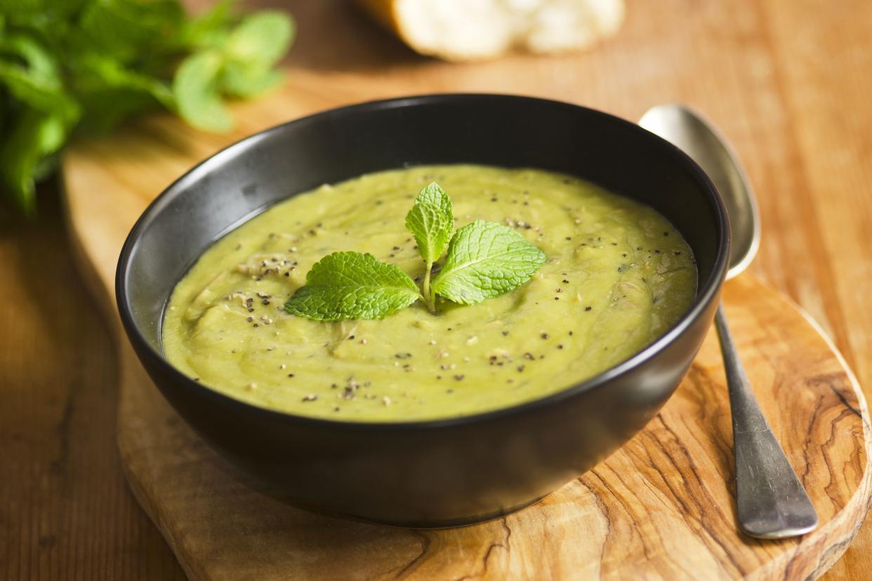 Easy pea soup in a dark brown ceramic bowl on an olive wood serving board with a spoon, on a wooden table with ingredients blurred in the background