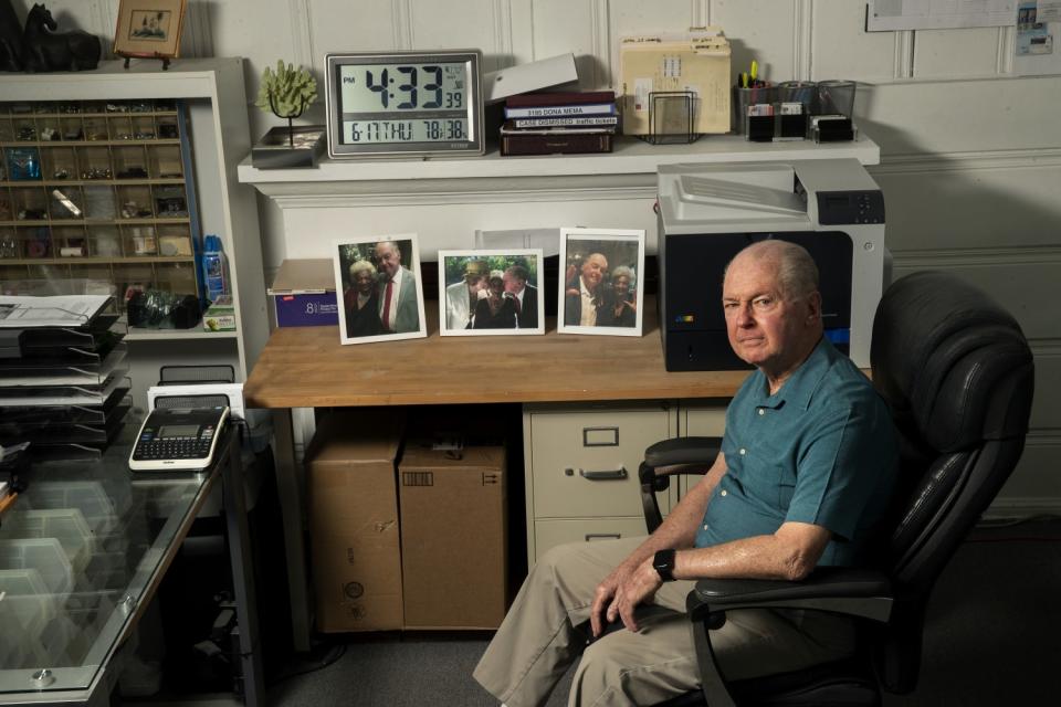 A man sits in an office chair in an office. On his desks are photos of himself and a friend.