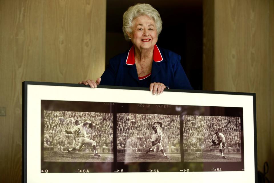 A woman in white hair holds a frame with three photos of a pitcher pitching.