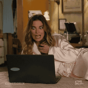 Annie Murphy, wearing a robe, is lying on a bed with a shocked expression while looking at a laptop screen in a scene from "Schitt's Creek" and says, "Yum!"