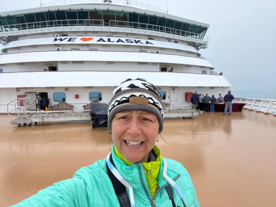 rebecca posing on a cruise ship in front of a "we heart alaska" sign wearing a hat and layers