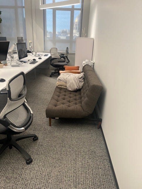 A sofa with bed sheets and pillows is on a floor with multiple desks.