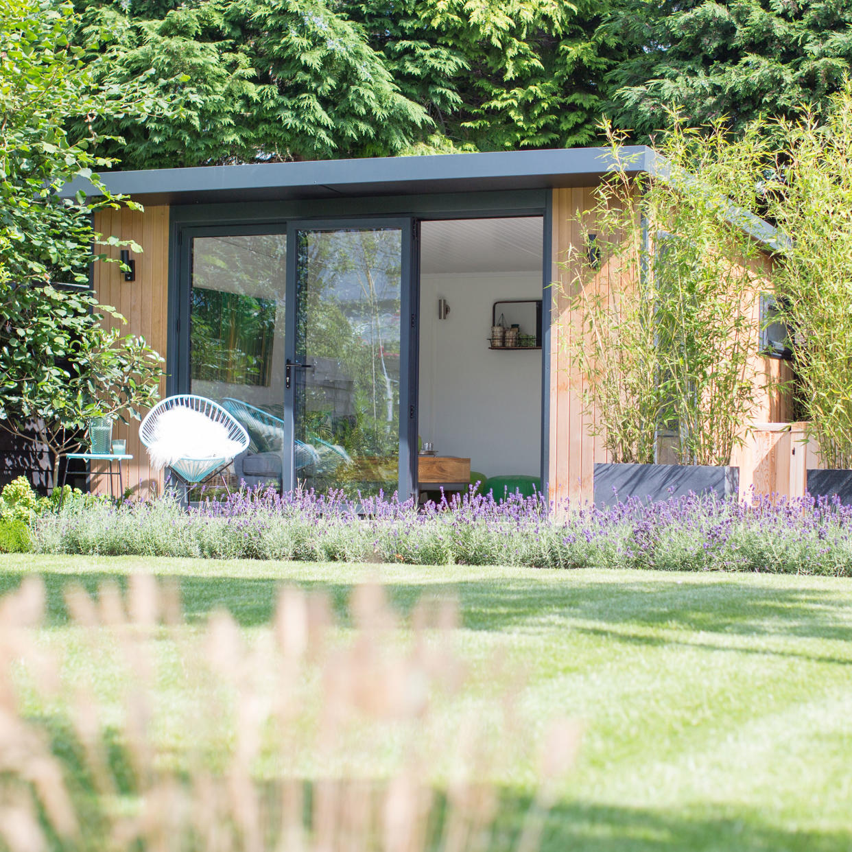  A garden summerhouse with sliding glass door, white walls, and green decorative accents, surrounded by plants. 