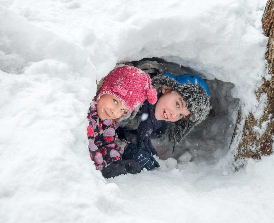 two children wearing toques and winter jackets playing inside snow fort outside 