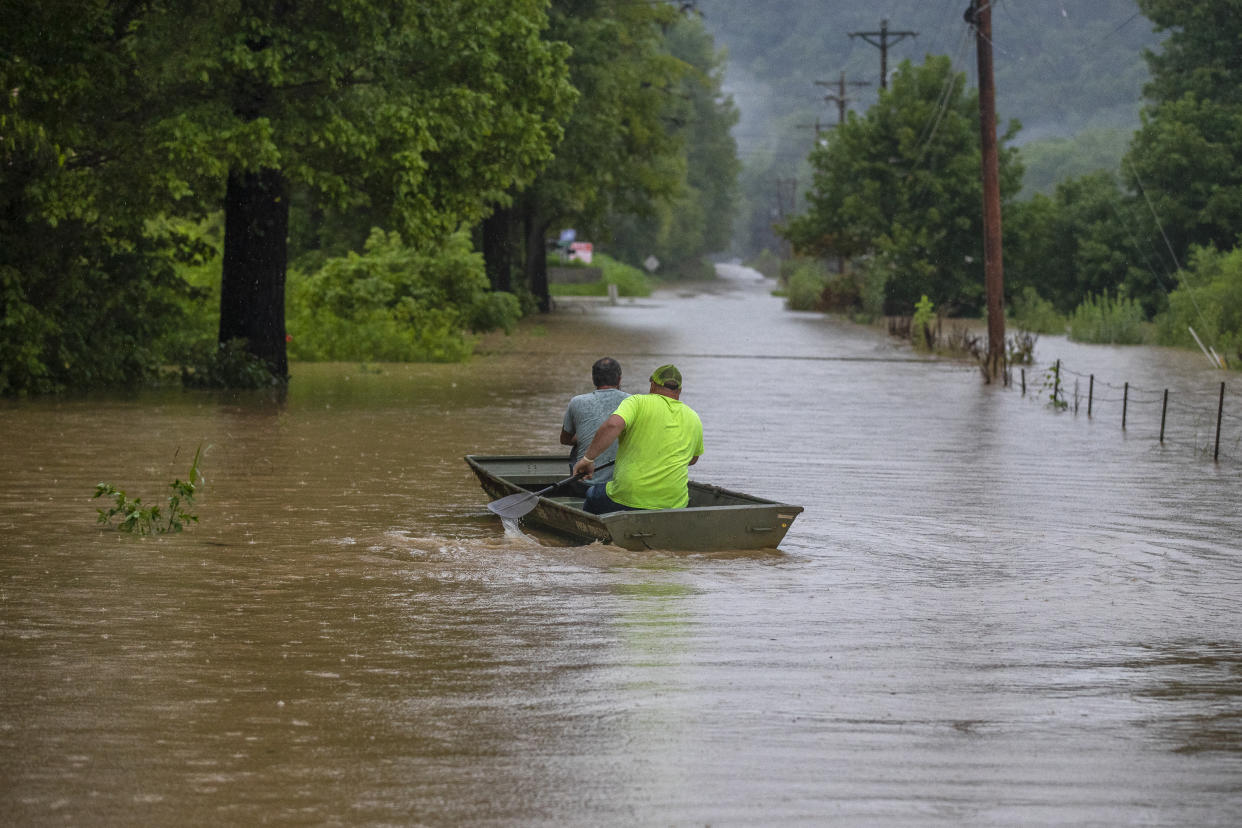 Two men paddle in a shallow boat along a flooded road.