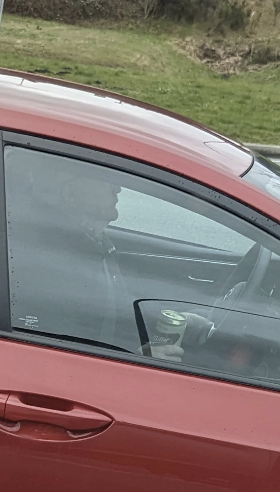 A guy driving a car with their left hand with a can of Miller in their right hand