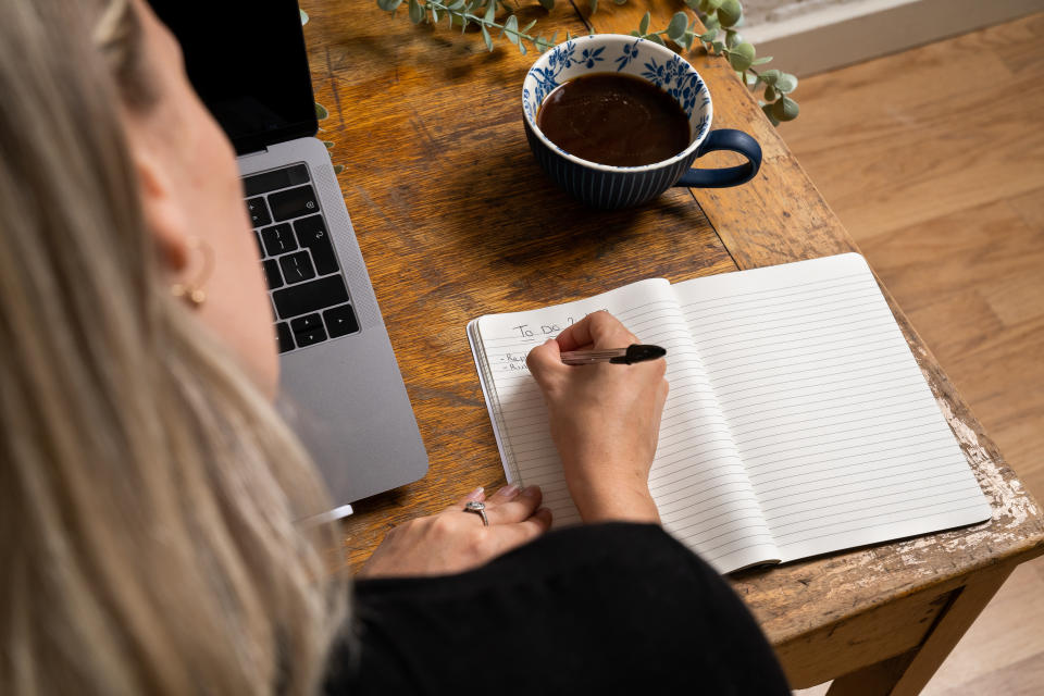Frau schreibt etwas auf einen Zettel, sie sitzt am Schreibtisch neben einem Laptop und einer Tasse Kaffee