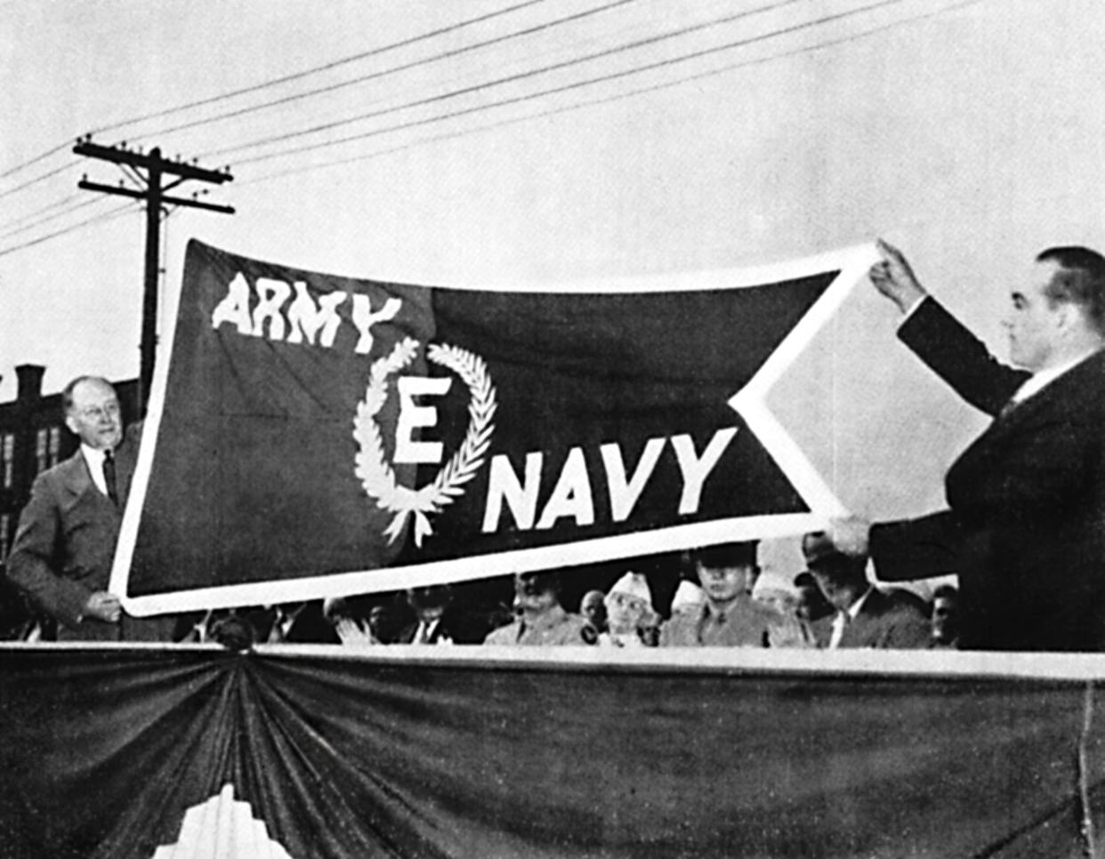 On August 18, 1942, Savage Arms in Utica received from the US War Department the Army - Navy “E” Flag for high achievement in the production of equipment for World War II, which was raging at the time. Its thousands of employees also received “E” lapel pins. Exhibiting the flag in a plant ceremony were Fred Hickey, left, the company president and Bob Havens, who represented the employees. Among the items Savage made were .50 caliber Browning aircraft machine guns and Thompson submachine guns. The plant—recently razed—was on Turner Street in the city’s east end and sat on 32 acres. Its parking lot accommodated 2,500 vehicles.