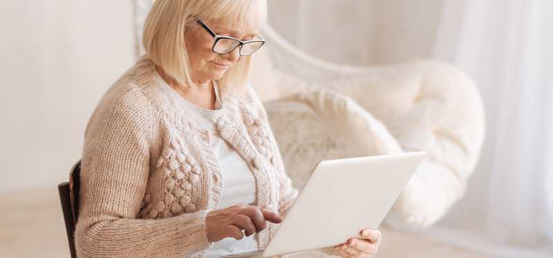 An elderly woman sits in a chair and uses a tablet.