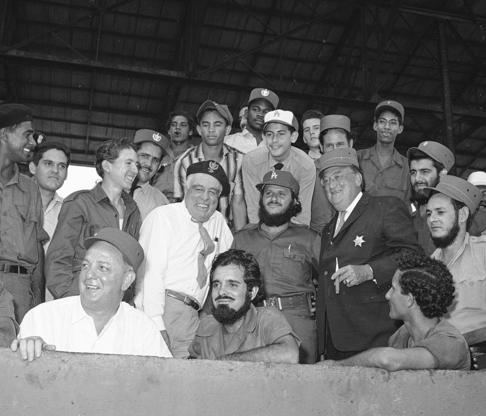 FILE - In this March 21, 1959, file photo, a Cuban baseball fan, center,  wears Los Angeles Dodgers' owner Walter O'Malley's cap while O'Malley sports a rebel's hat and a sheriff's badge during an exhibition baseball game between the Dodgers and Cincinnati Redlegs in Havana, Cuba. At left center, Dodgers director Bud Holman grins wearing an army captain's beret.  Reds' general manager Gabe Paul, seated at lower left, also tries out a rebel officer's hat. Others are unidentified. The game would be the last between major league teams in Cuba for the next four decades. (AP Photo/File)