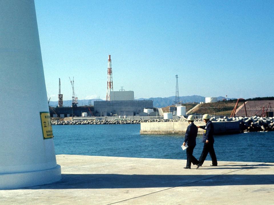 Two men in construction hats walk on a concrete pier with the Fukushima power plant in the background.