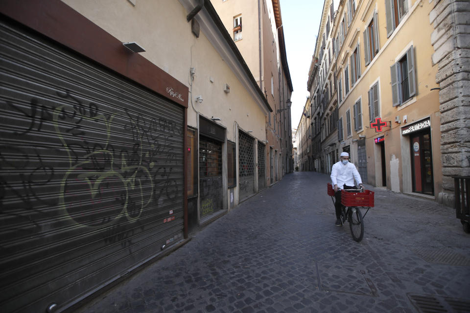 FILE - In this March 15, 2021, file photo, a man wearing a face mask to curb the spread of COVID-19, pedals past closed shops in downtown Rome. Optimism is spreading in the U.S. as COVID-19 deaths plummet and states ease restrictions and open vaccinations to younger adults. But across Europe, dread is setting in with another wave of infections that is closing schools and cafes and bringing new lockdowns. (AP Photo/Alessandra Tarantino, File)