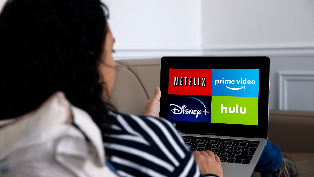 A woman looking at a laptop screen showing the logos of Netflix, Amazon Prime, Disney Plus and Hulu.