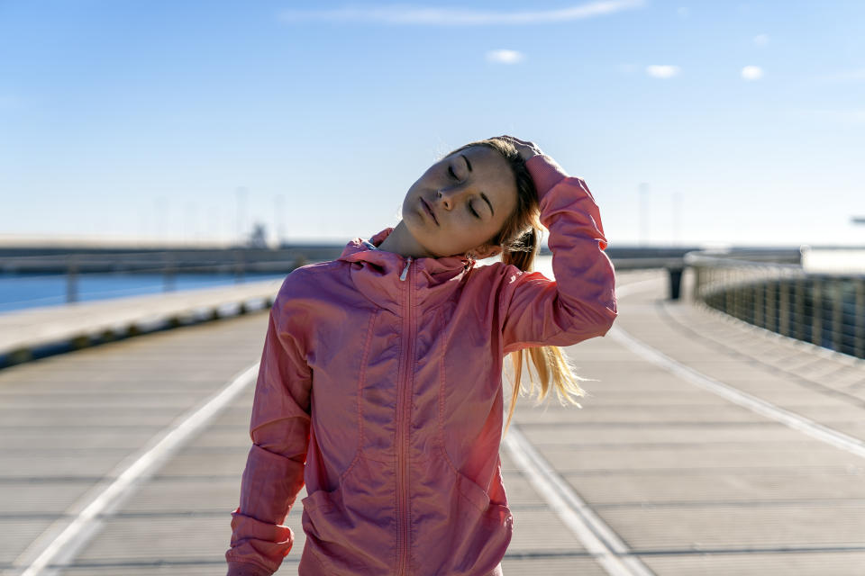 A woman wearing a pink running jacket stands on a pier and places a hand on her head to stretch the side of her neck