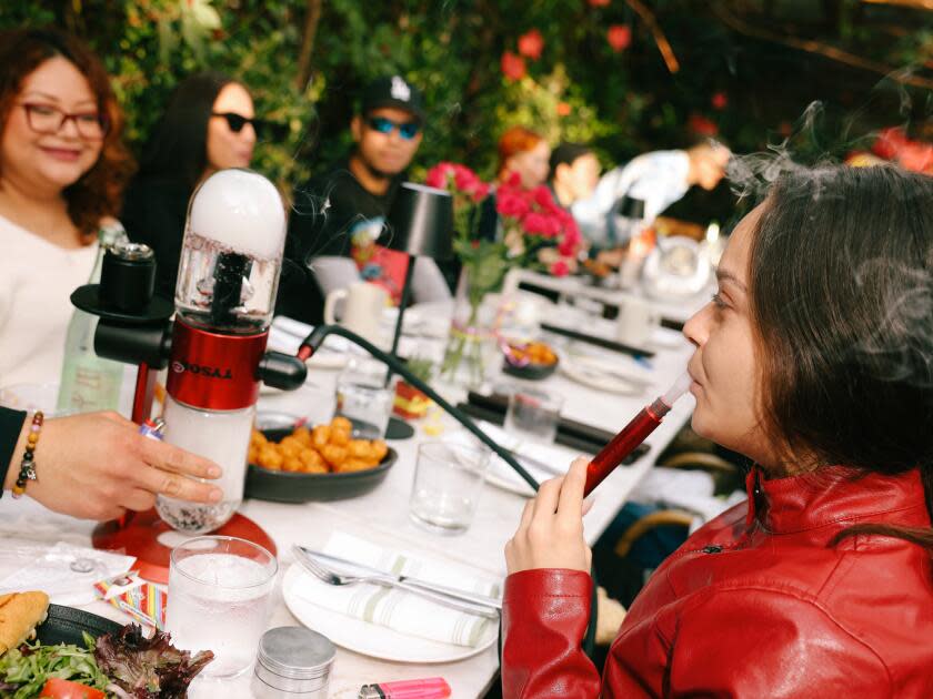 A woman sits at a long table with several other people. She holds a hose next to her mouth with smoke coming out.