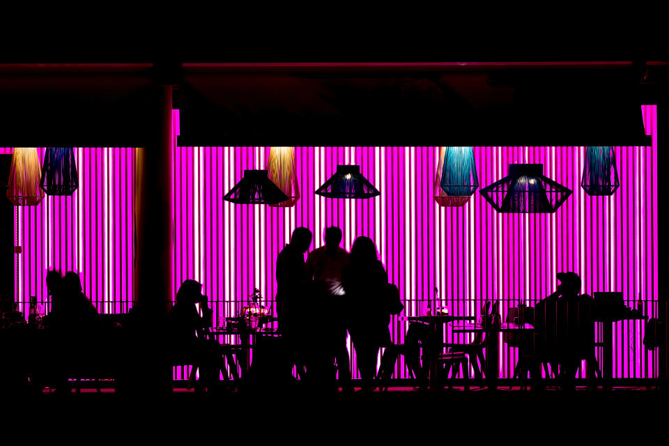 People sitting and standing at restaurant tables outside, silhouetted against a neon-striped background