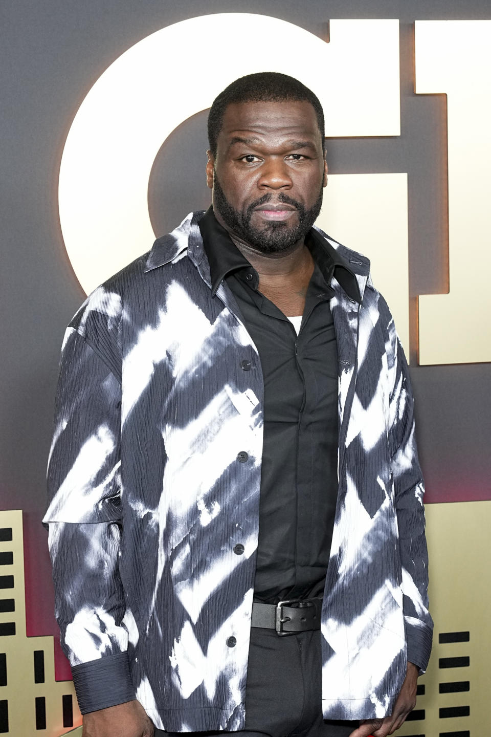50 Cent in a tie-dye patterned jacket over a black shirt, standing in front of a promotional background