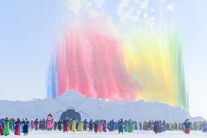 A large group of people in colorful clothing stand in front of a snow castle as colorful fireworks explode in the sky above