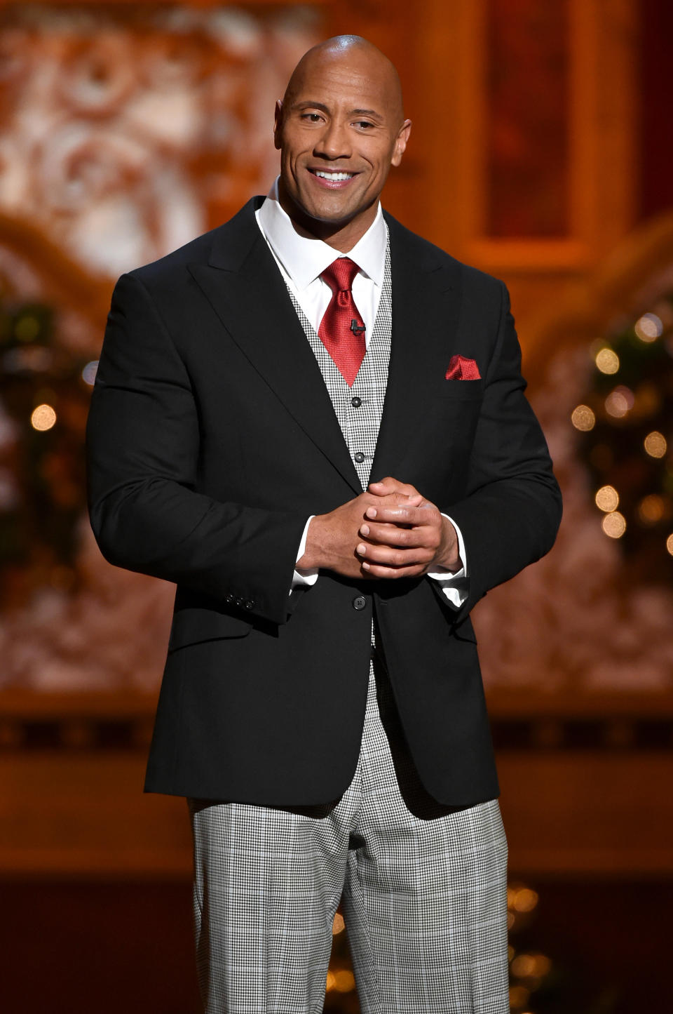 Dwayne "The Rock" Johnson is wearing a checkered three-piece suit with a red tie and pocket square, smiling at the camera while standing in front of a festive background