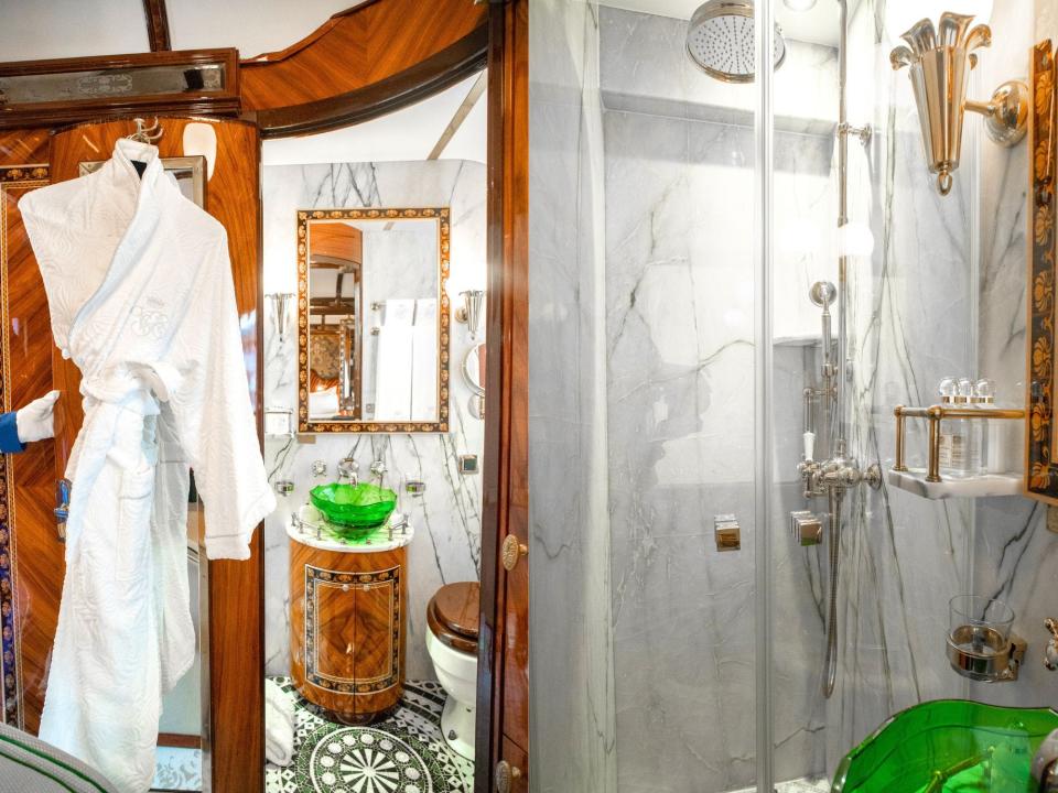 Left: A wooden door opens to reveal a marble bathroom, white bathrobes hanging on the left. Right: A silver shower head behind a glass door in a bathroom with marble walls