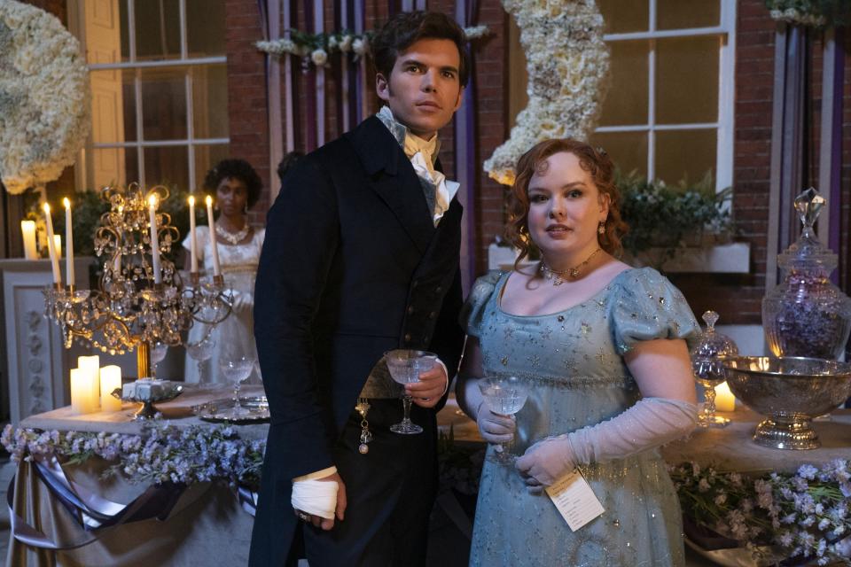 Luke Newton and Nicola Coughlan in 19th-century formal attire, holding champagne glasses at an elegant candlelit party