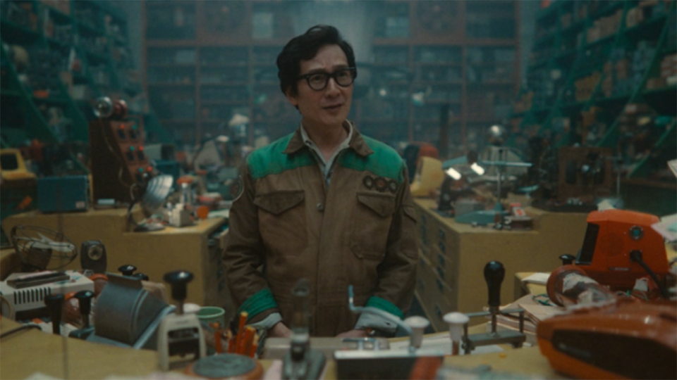 a man in a denim jumpsuit stands in a crowded library full of books and gadgets