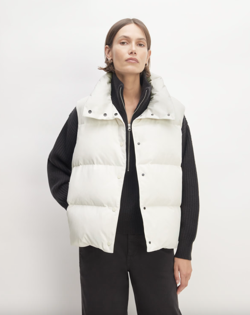 model wearing black pants, black sweater and white puffer vest, The Re:Down Vest in bone (photo via Everlane)