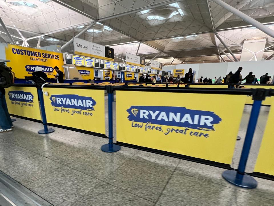 Yellow signs with the Ryanair logo as people line up for the check in desk and bag drop at Stansted Airport
