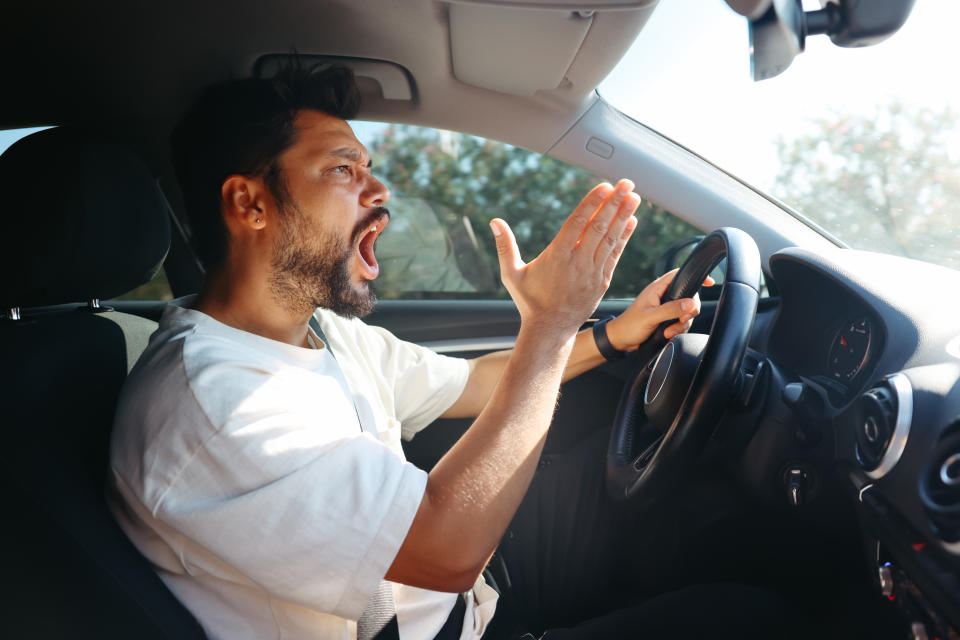 A man with a beard, wearing a white shirt, is in the driver's seat of a car, expressing frustration. The background shows a blurred outdoor setting