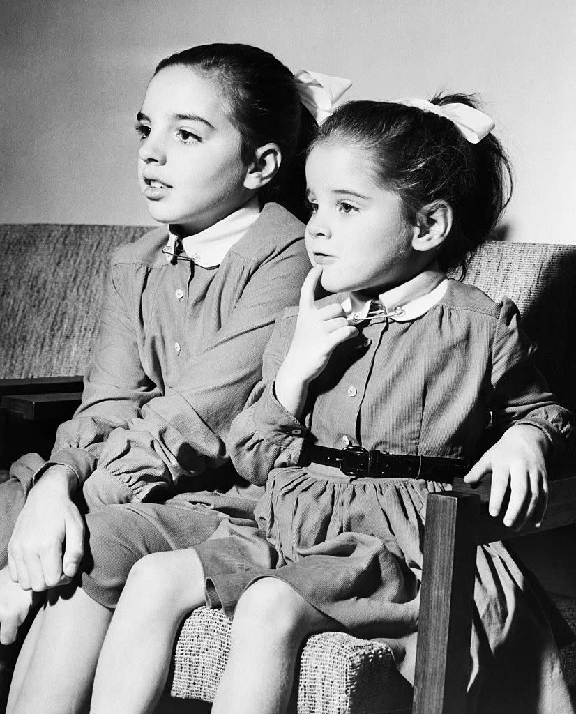 Two young girls, one with her finger on her chin, are sitting on a couch wearing matching long-sleeved dresses