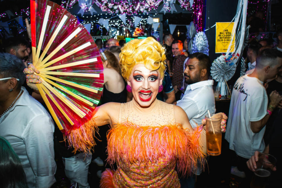 A drag queen with yellow hair and an orange dress holds a red fan and smiles