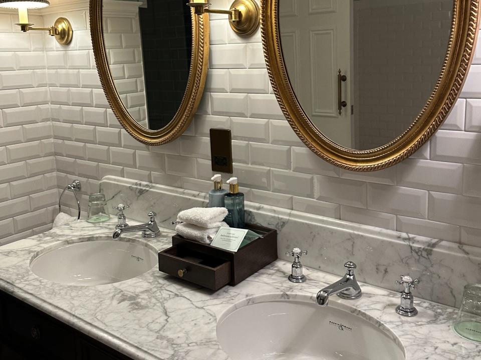 A view of double marble sinks in a hotel bathroom. There is a gold oval mirror above each