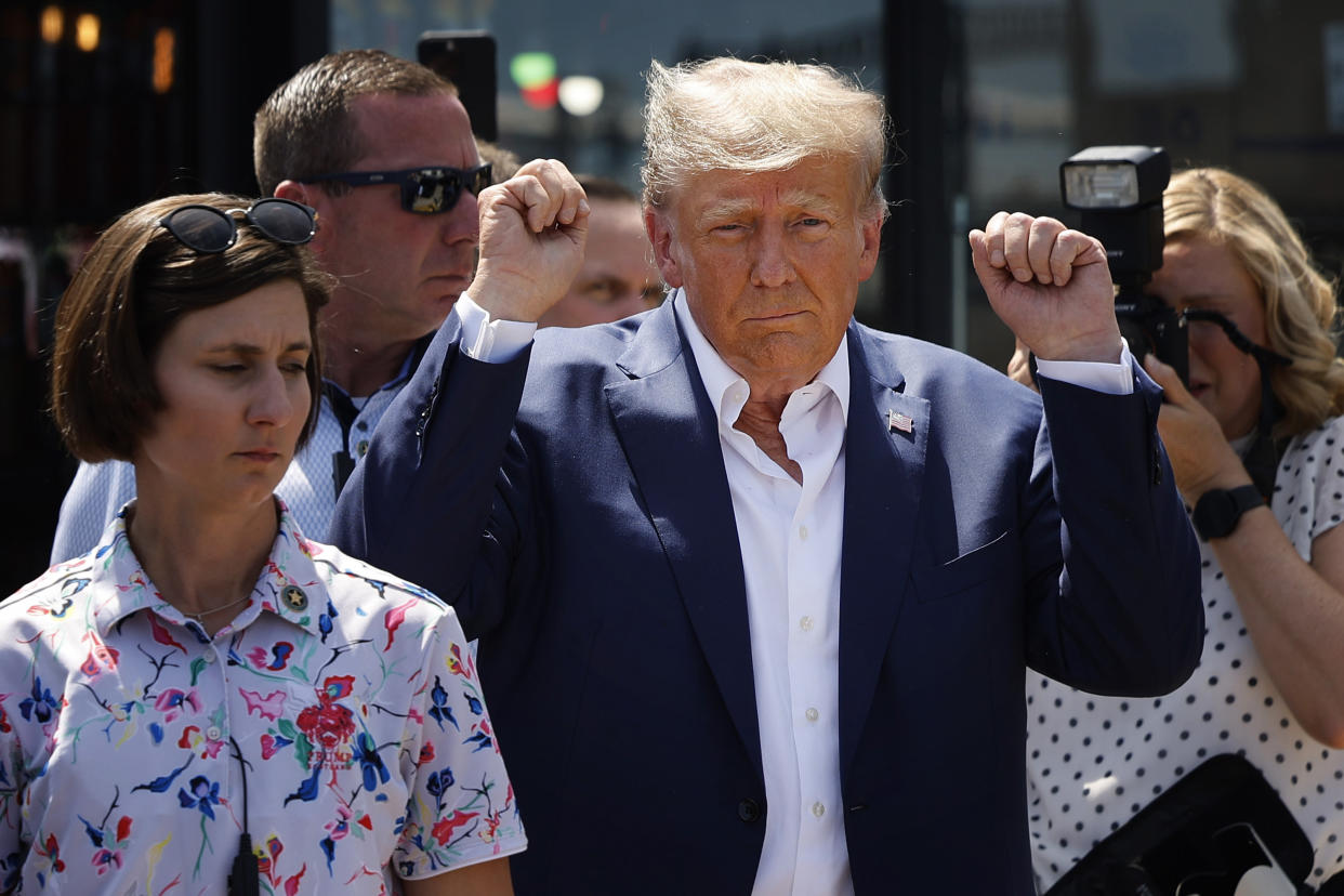 Donald Trump, surrounded by people wearing summer clothes, raises two fists unconvincingly in the air.