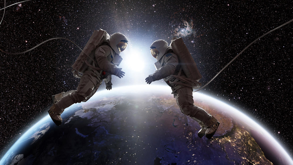  Two astronauts in full spacesuits with backpacks on a space walk with tethers, facing each other with hands out. They are floating in the front of Planet Earth as the sun rises between them. 