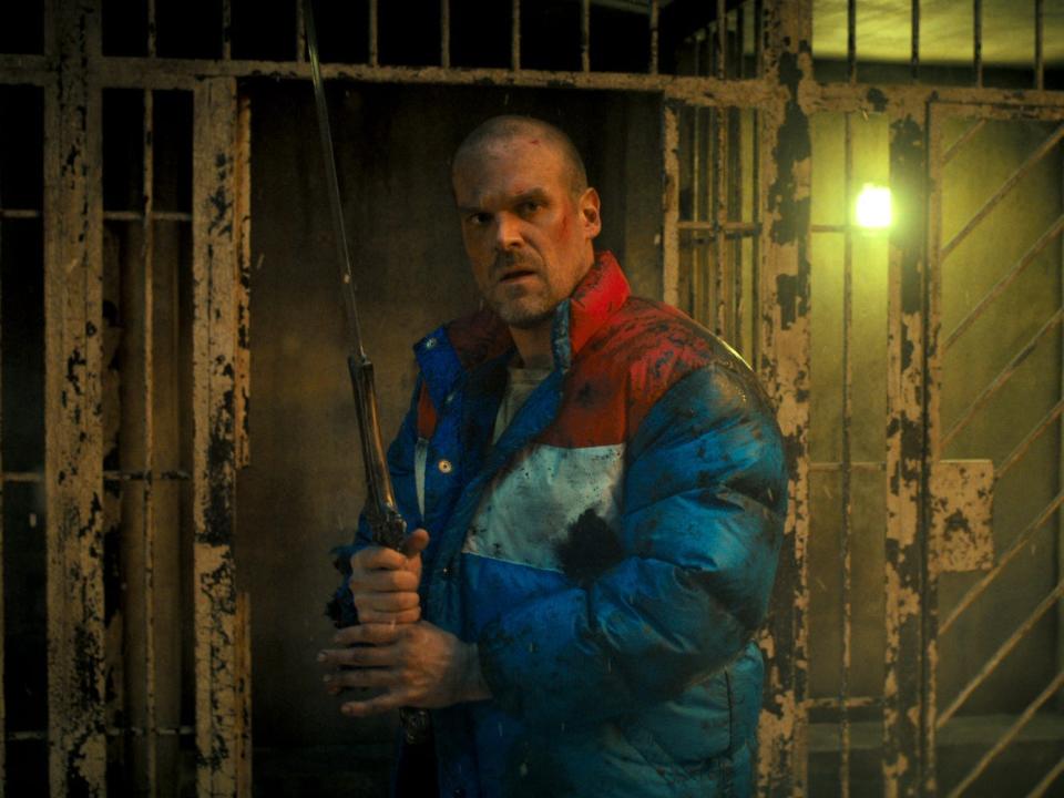 A man with a shaved head and wearing a puffy jacket holds up a metal sword.