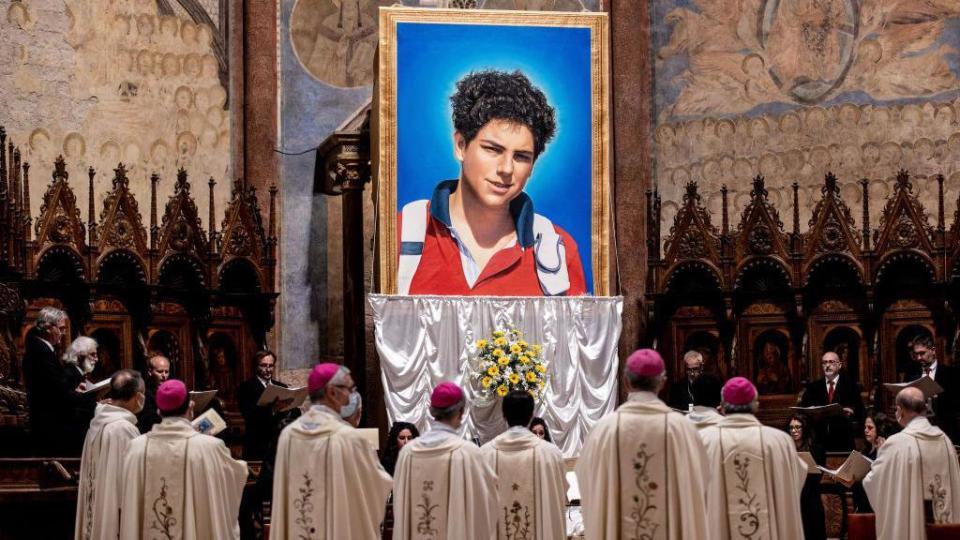 Un tapiz de Carlo Acutis, un adolescente, y personas con atuendos religiosos mirando.