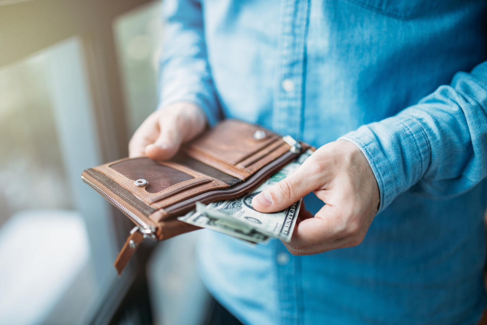 A person in a denim shirt is holding an open wallet and removing dollar bills