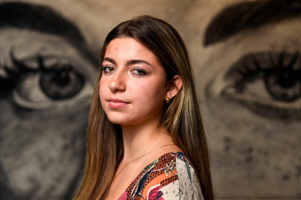 Anna Castro Spratt, 16, Greenville's first Youth Poet Laureate, poses for a portrait in front of a painting by fellow student, Hannah Brewer, inside the Fine Arts Center at Wade Hampton High School on Wednesday, May 3, 2023.